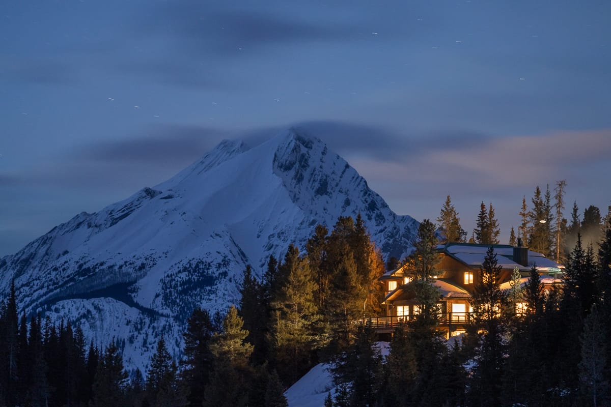 view of mount engadine lodge at night, the lights in the windows are glowing and there is a snow covered mountain in the background