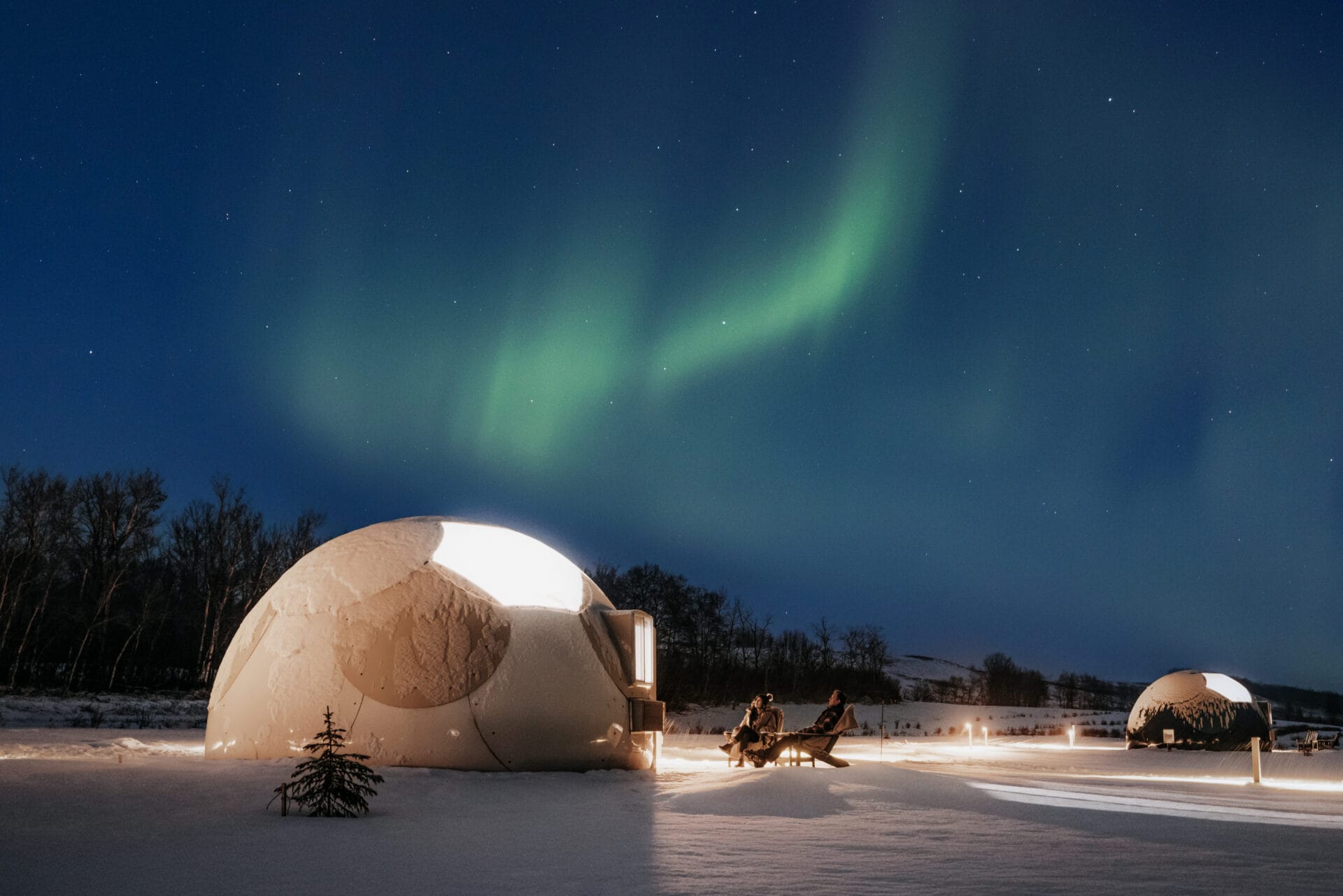 a couple stare up at the northern lights while sitting outside their dome glamping tent at Metis crossing