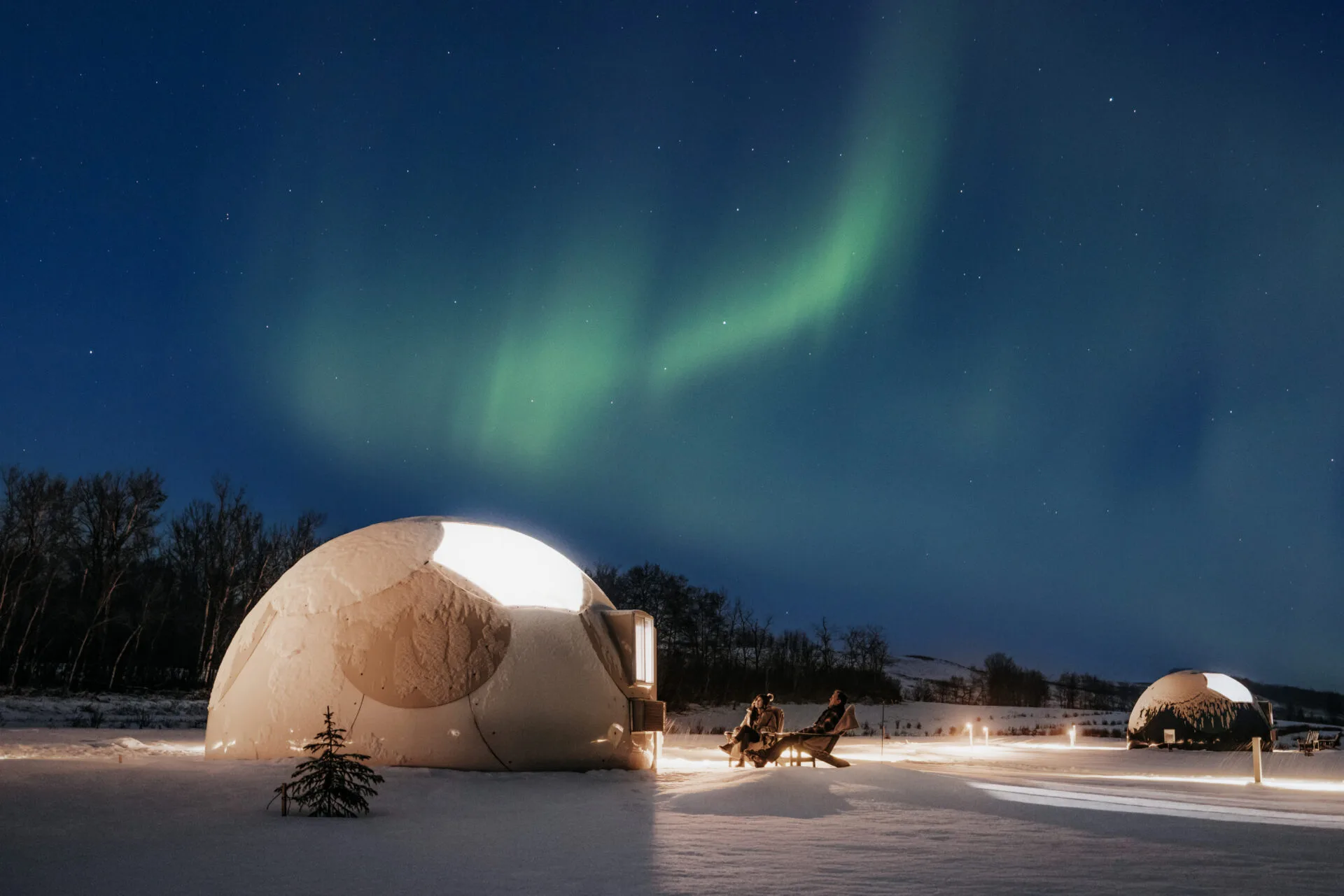 a couple stare up at the northern lights while sitting outside their dome glamping tent at Metis crossing