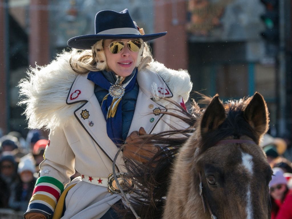 a cowgirl rides a horse, she is wearing a white coat, cowgirl hat and sunglasses