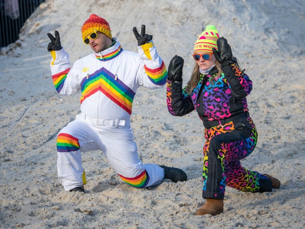 two people kneel in the snow while wearing their bright coloured retro ski suits