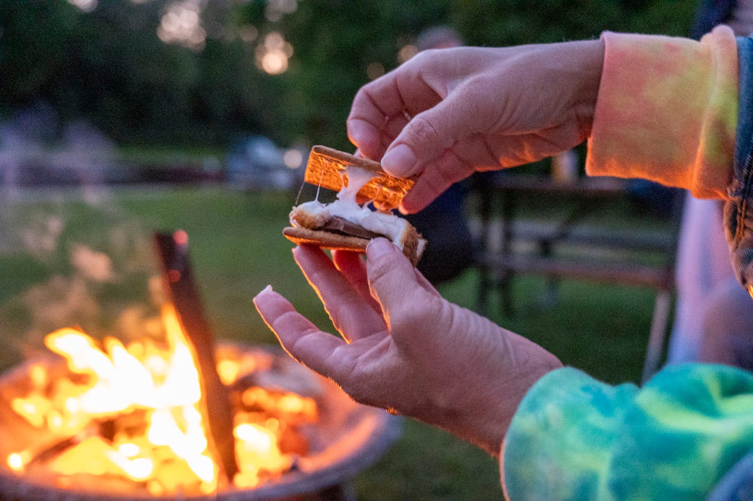 close up of two hands opening a gooey s'more, a campfire is in the background.