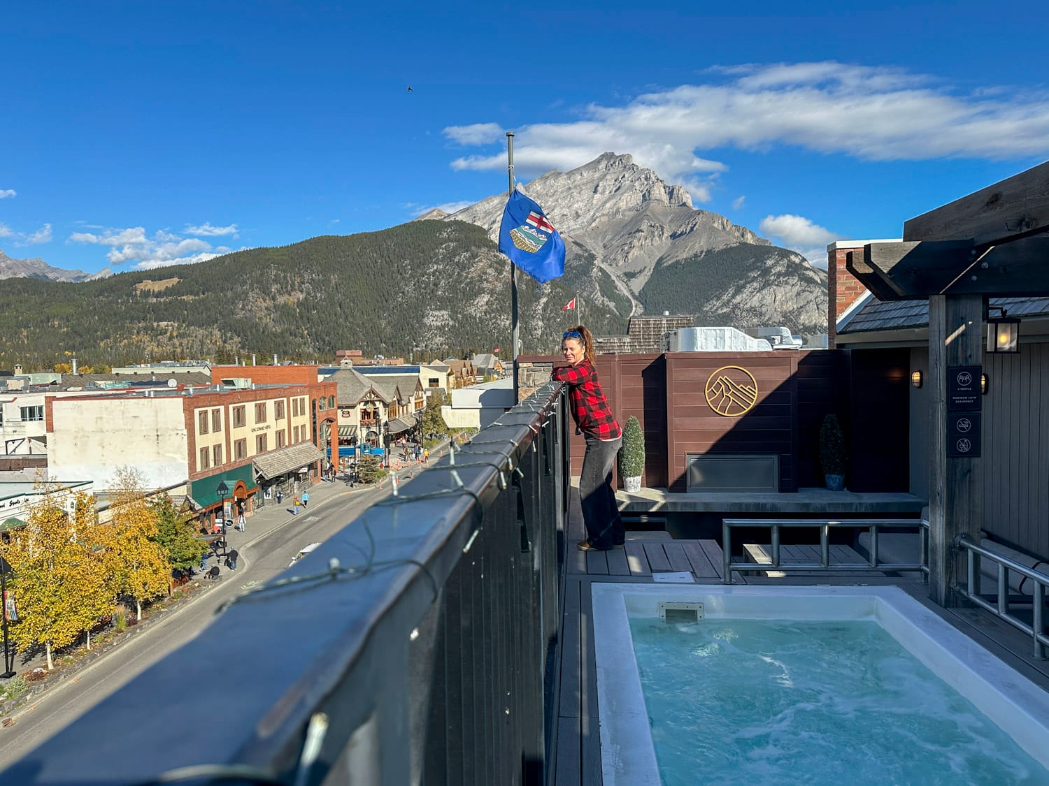 woman stands on the top deck of the mount royal hotel, beside their outdoor hot tubs, the town of banff and mountain are in the background