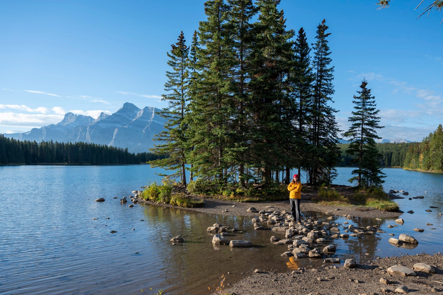 woman wearing a bright yellow coat stands on the shore of 2 jack lake in banff national park