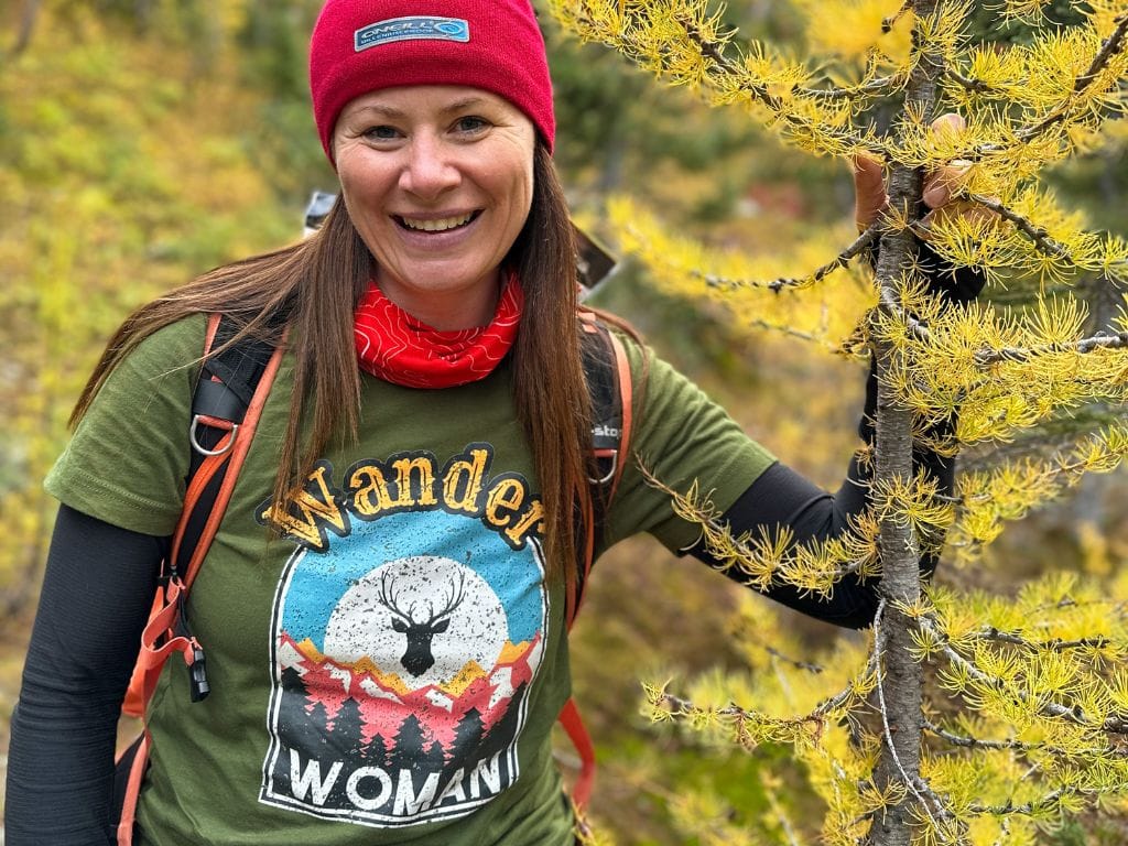 woman wearing a red toque and a green shirt that reads "wander woman" standing next to a yellow larch tree