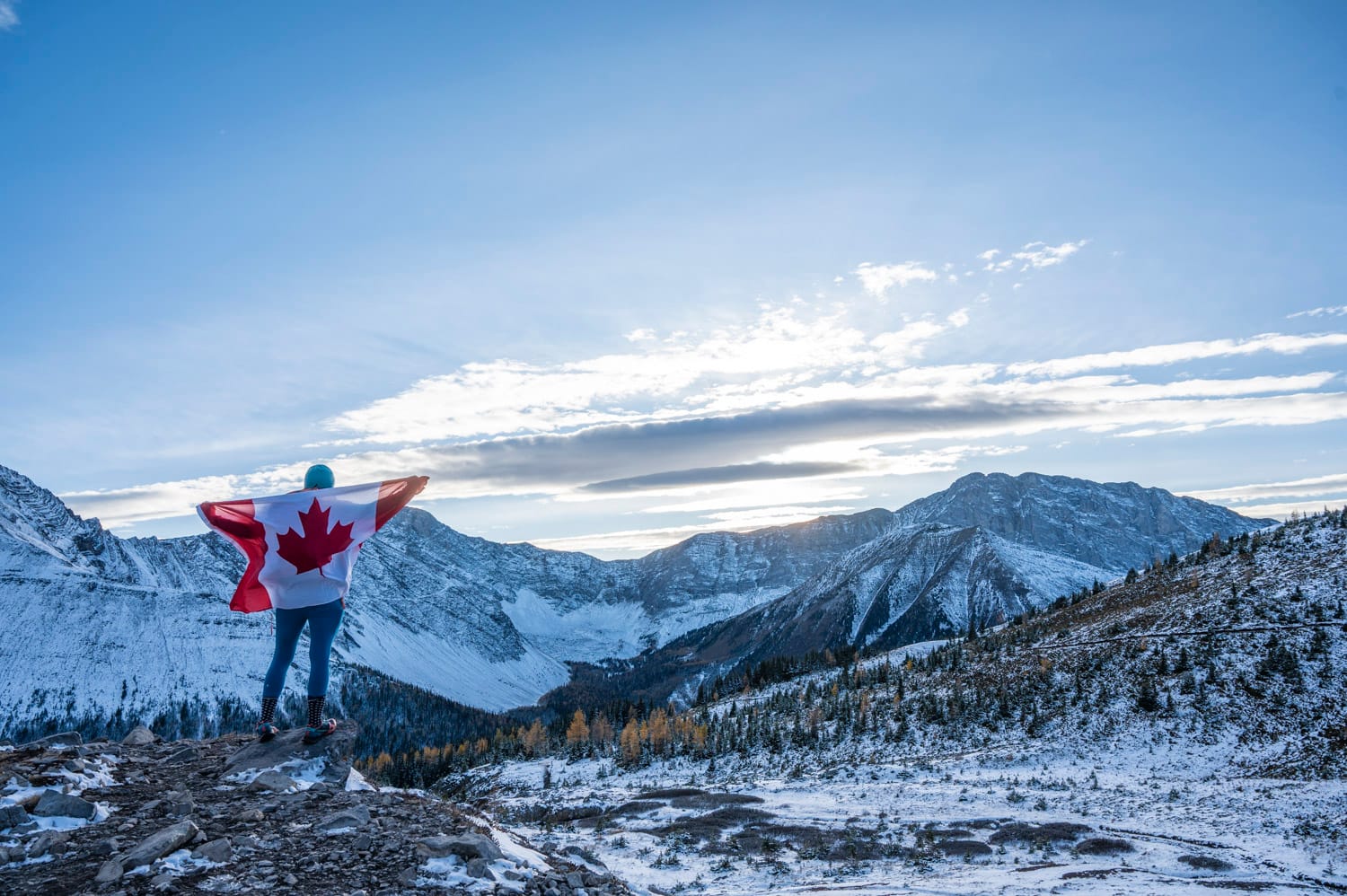 a woman stands with the canada flag at her back, she is looking out at snow covered mountains and yellow larch trees