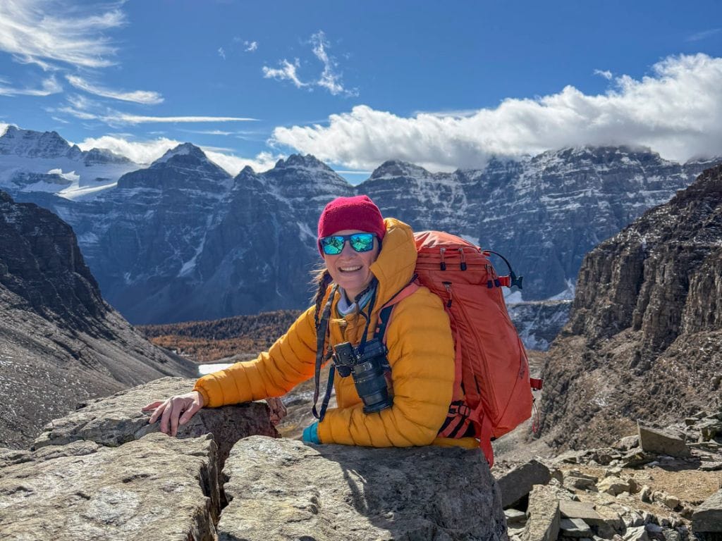 a woman wearing winter gear and sunglasses smiles at the camera from the top of sentinel pass hike during larch season banff