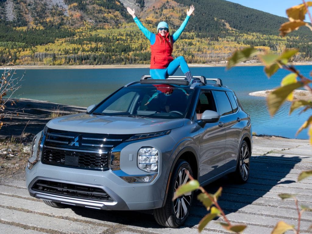 woman sits on the top of a Mitsubishi outlander through the sunroof, mountains and a lake are in the background