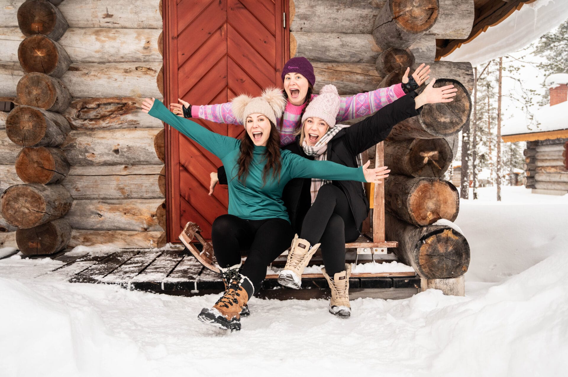 The Solo Travel Collection - Three women in winter clothing, smiling and posing with outstretched arms in front of a log cabin in Finland