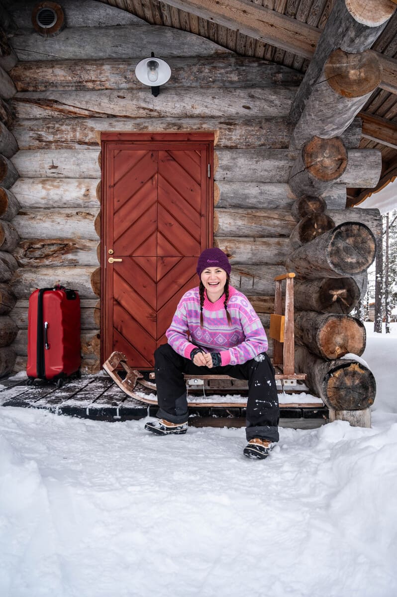 A woman in winter clothes sitting on a sled in front of a log cabin, with luggage next to her