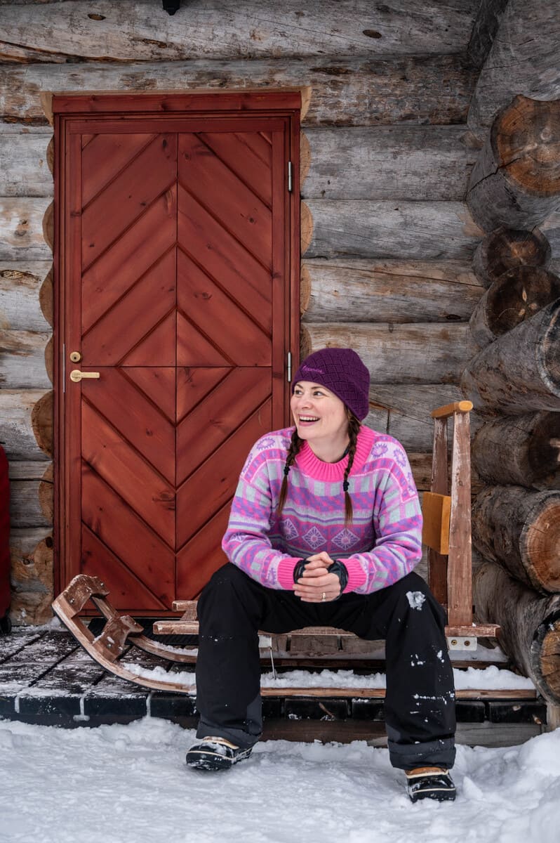 A woman in winter clothes sitting on a sled in front of a log cabin