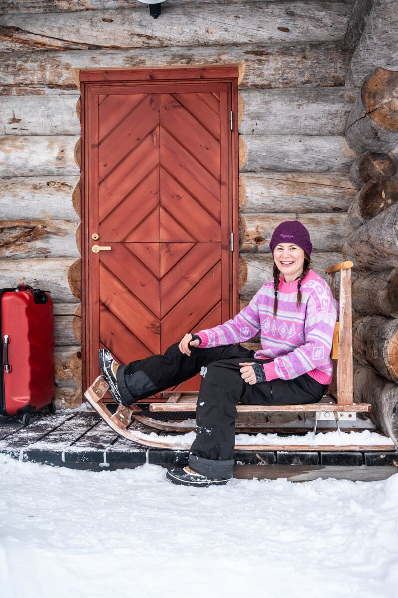 A woman in winter clothes sitting on a sled in front of a log cabin, with luggage next to her