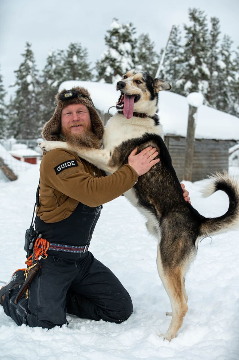 Man in winter gear kneeling in snow, smiling as he embraces a large, happy dog with its tongue out