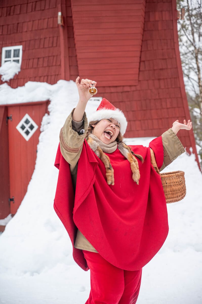 A joyful person in a Santa hat and red cape stands in snow, holding a bell and basket, exuding festive cheer
