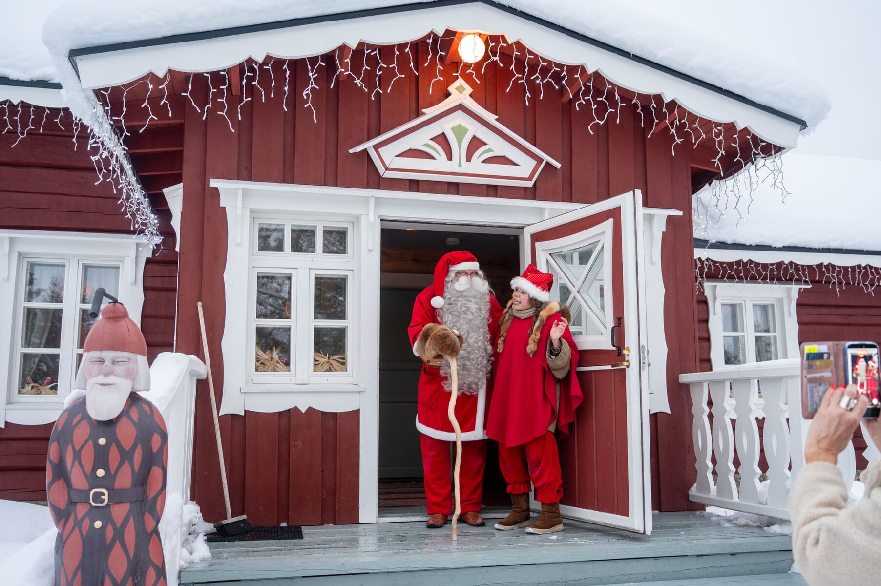 A cheerful person wearing a Santa hat and red cape standing next to Santa Claus