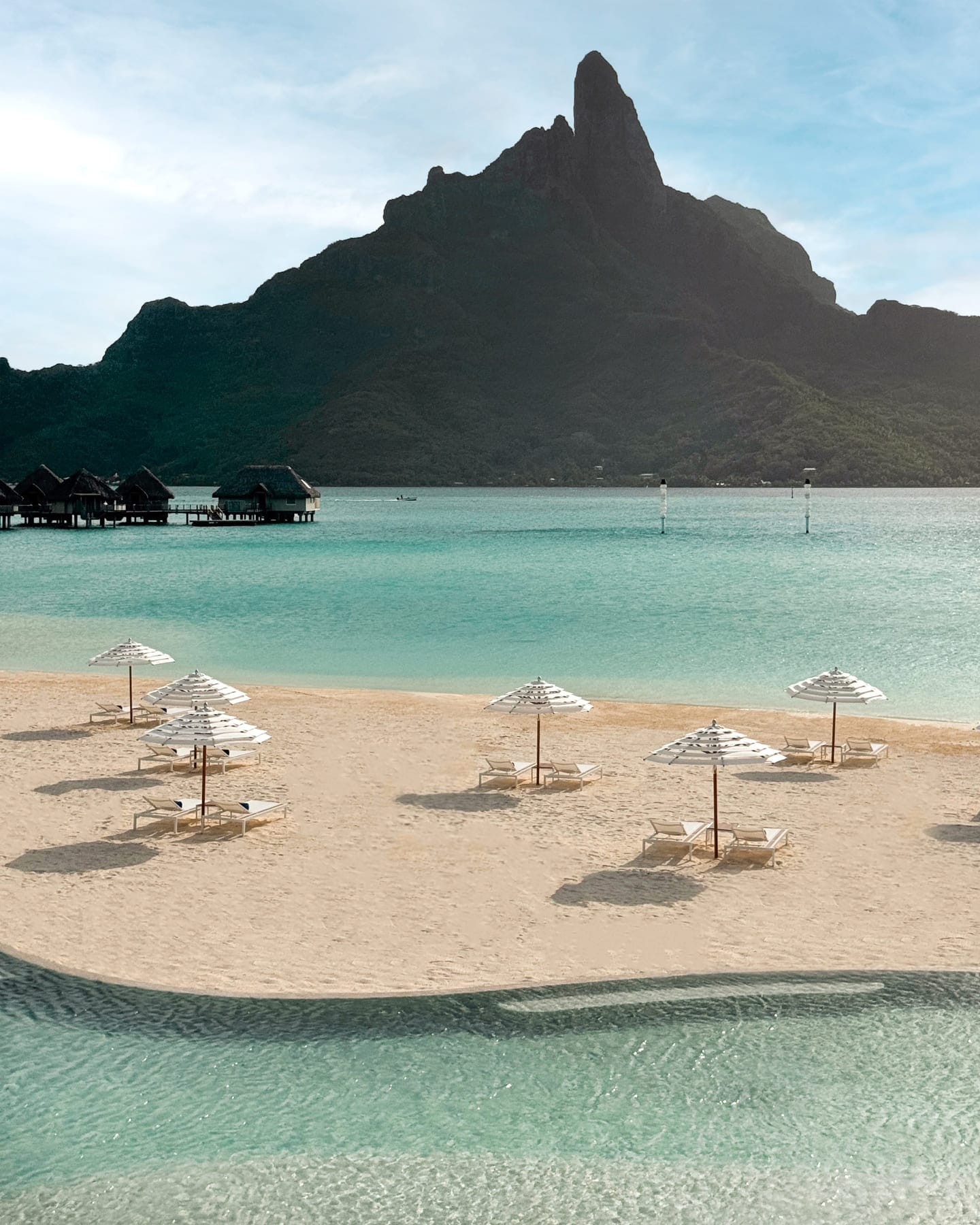 white sand beach with striped umbrellas, turquoise water, overwater bungalows, and a Mount Otemanu