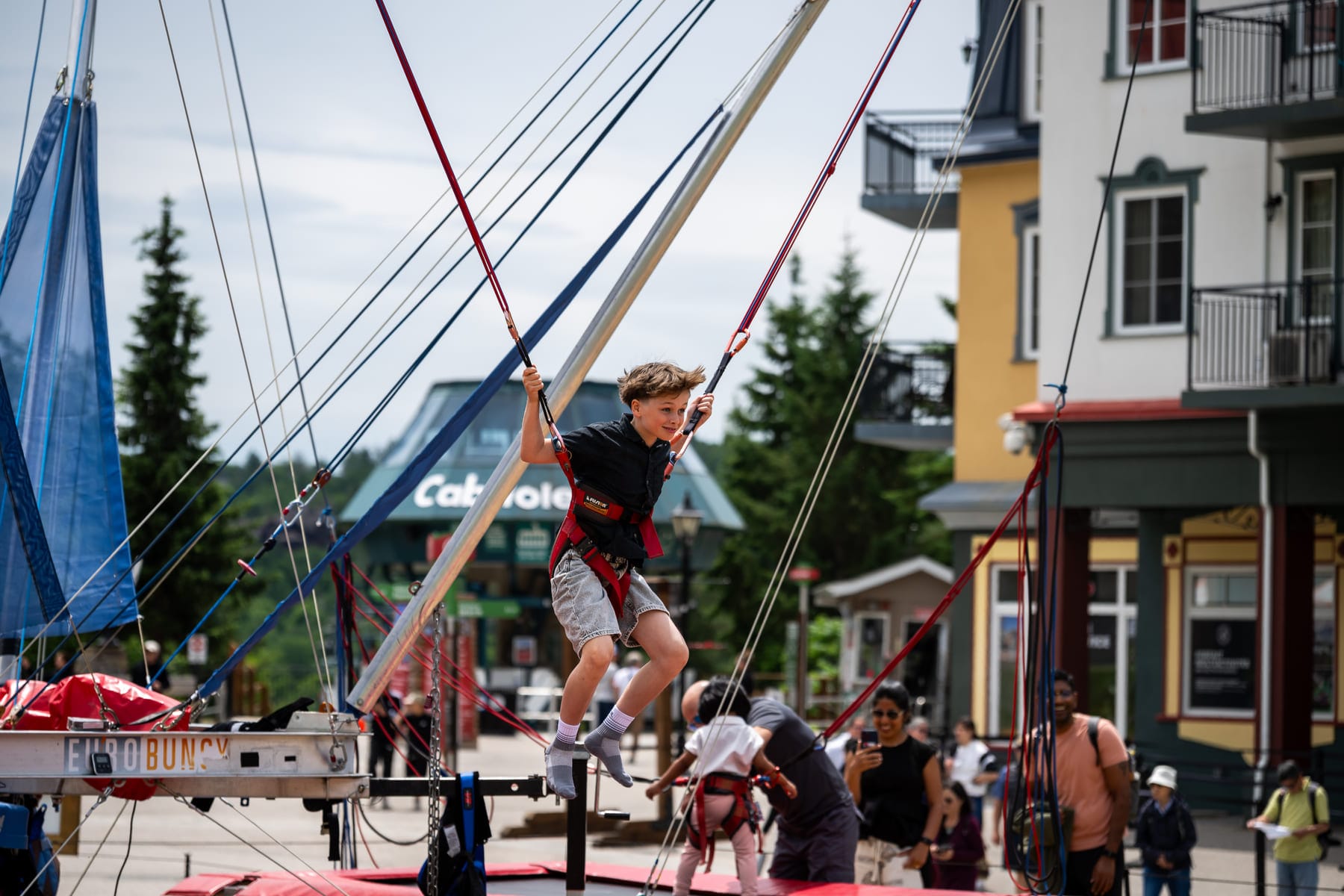 a boy attached to red harnesses and cords jumps on a Eurobungy trampoline with a happy expression; buildings and trees in background