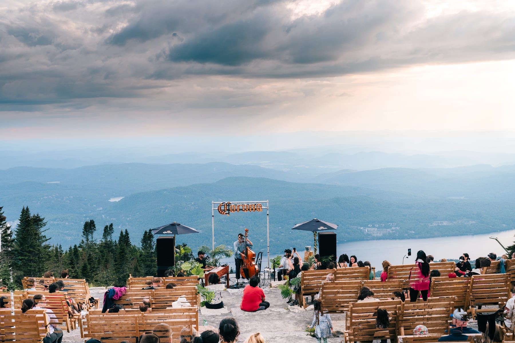 Outdoor concert on a mountain overlooking a lake and layered mountains with a band playing a string bass and keyboard, and attendees on wooden benches