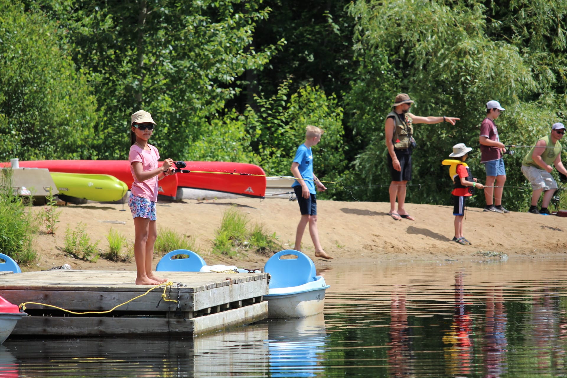 People fishing from a dock and shore with canoes, paddle boats, and green trees