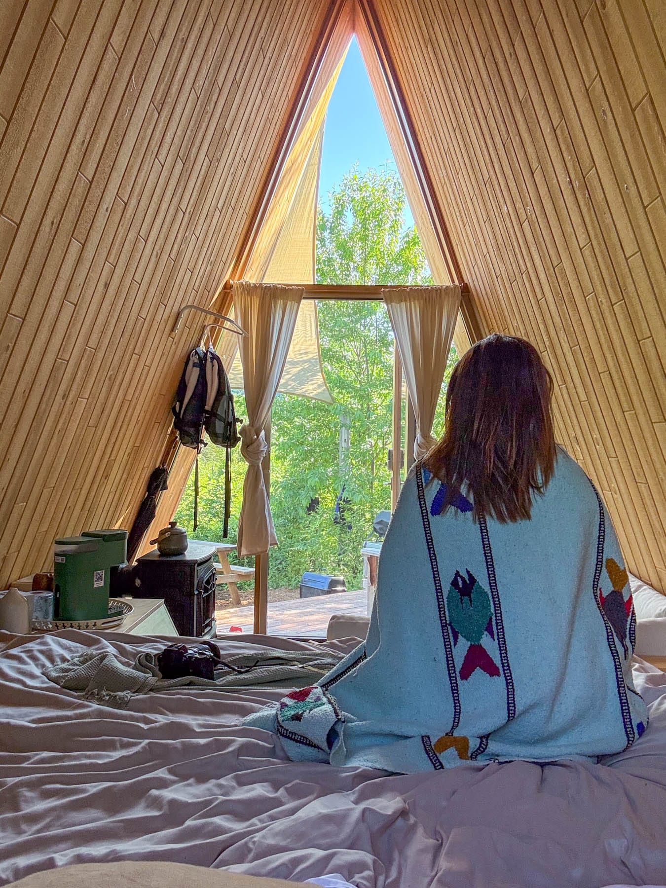 person in A-frame cabin, draped in a fish-embroidered blanket, looking out at a sunny green forest
