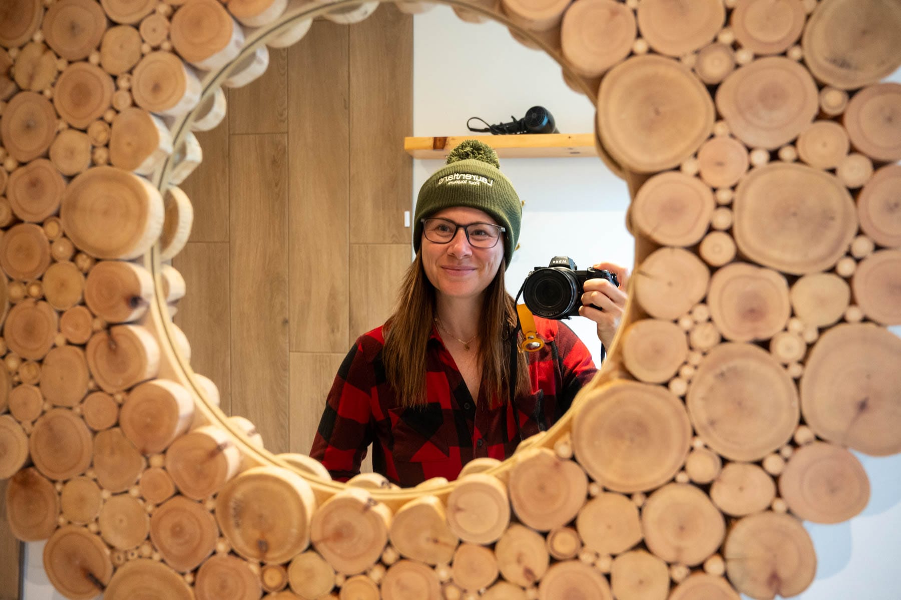 Jami Savage in plaid shirt and green beanie taking a mirror selfie with a Nikon camera in a round log-framed mirror