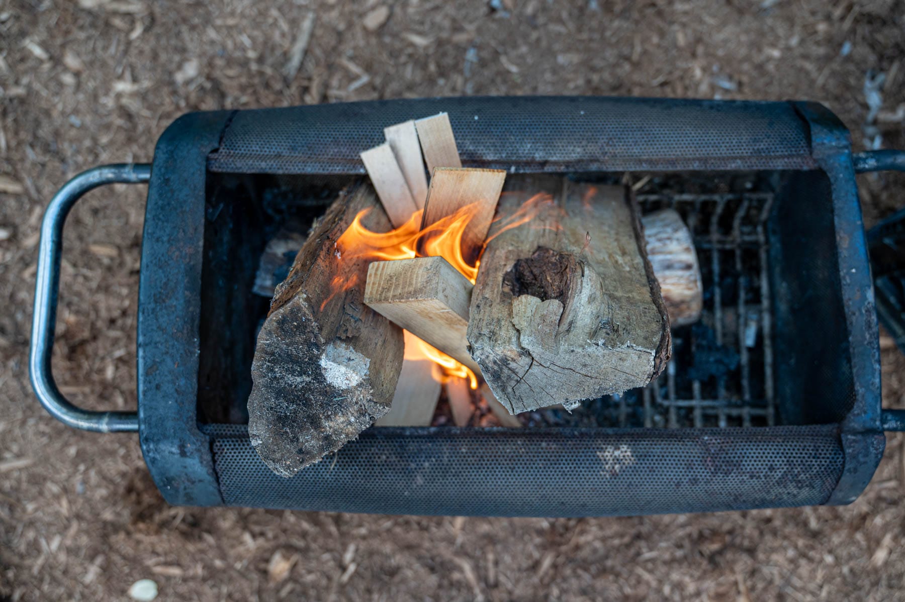 portable black metal grill with wood burning inside on grates, surrounded by wood chips