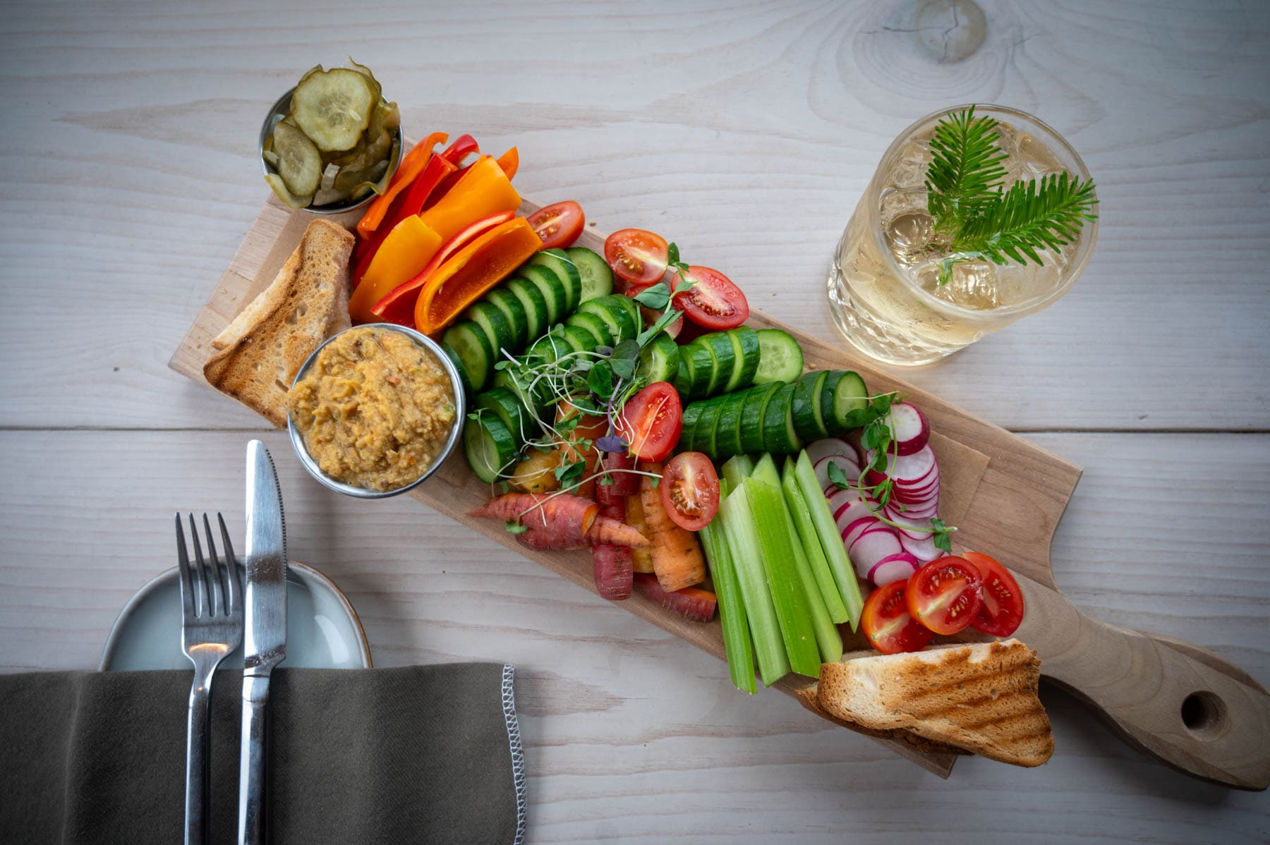 vegetable platter with various fresh produce, dips, and toasted bread on a wooden board, with a drink and cutlery
