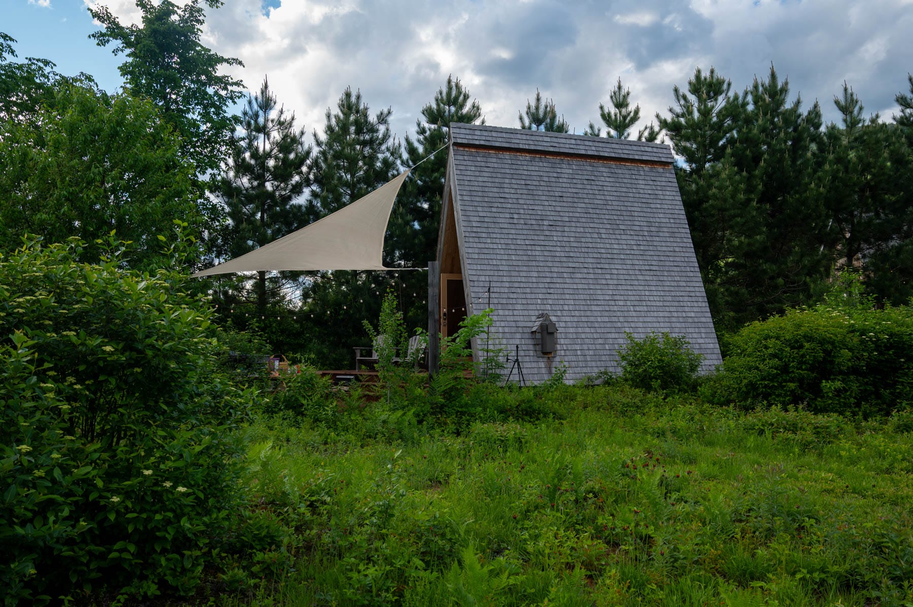 modern Farouche Tremblant A-frame cabin with gray shingles, surrounded by green foliage and pine trees, with a patio and sunshade
