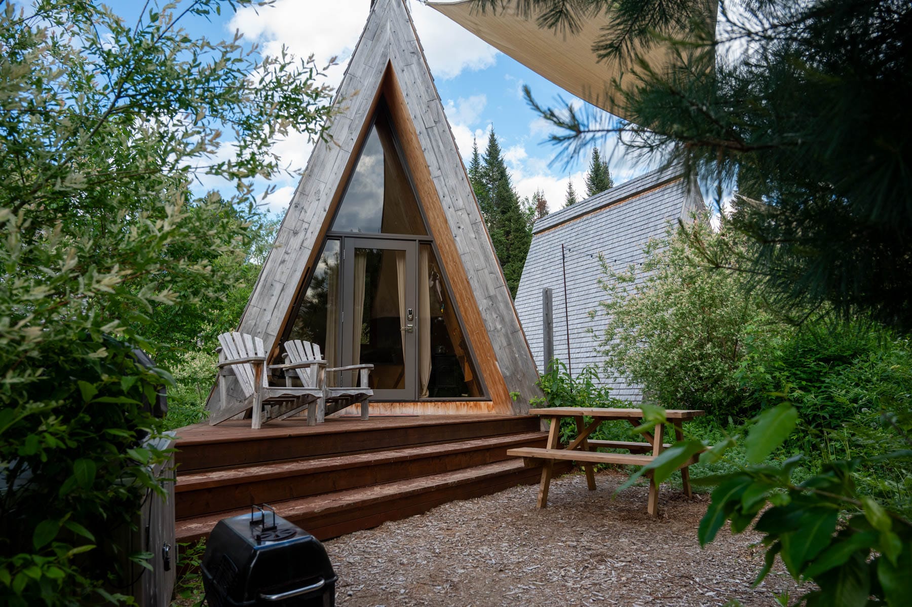 Farouche Tremblant A-frame cabin with a glass door, wooden deck, two Adirondack chairs, picnic table, and grill surrounded by greenery