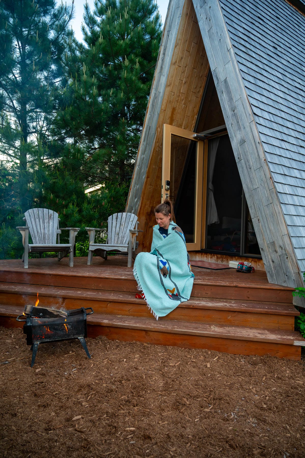 woman on A-frame cabin steps with fire pit, blue blanket, pine trees, Adirondack chairs; cozy cabin retreat