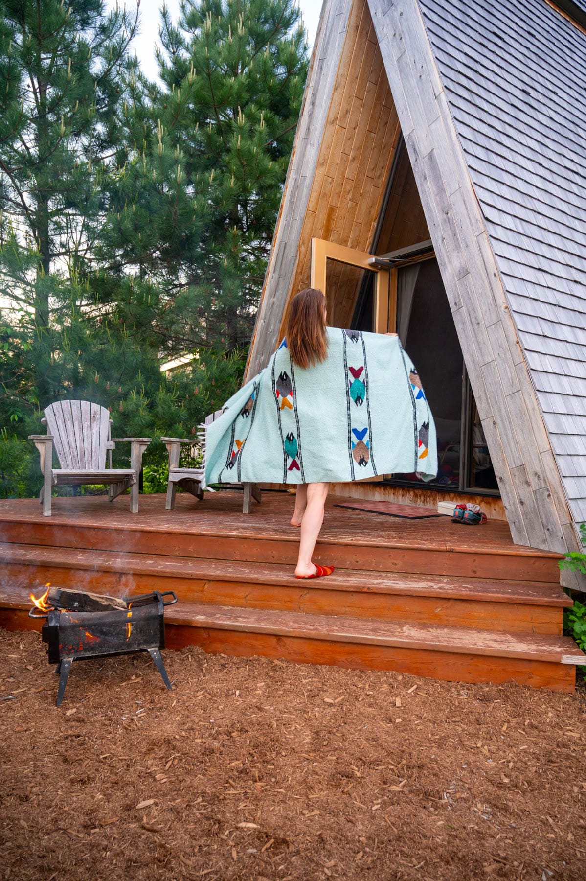 Jami Savage in a patterned blanket walking towards an A-frame cabin with a fire pit and Adirondack chairs nearby