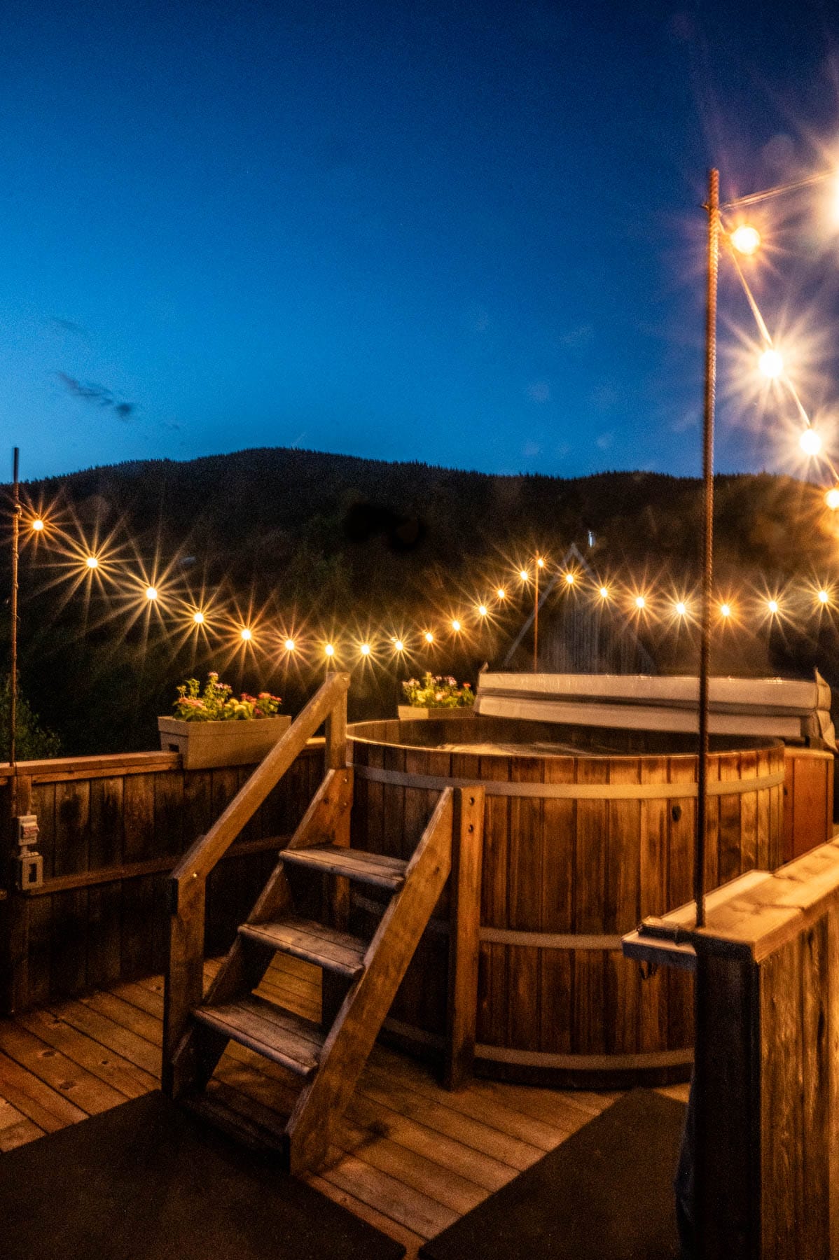 wooden hot tub on a deck with string lights at dusk, backed by a shadowy hillside and blue sky