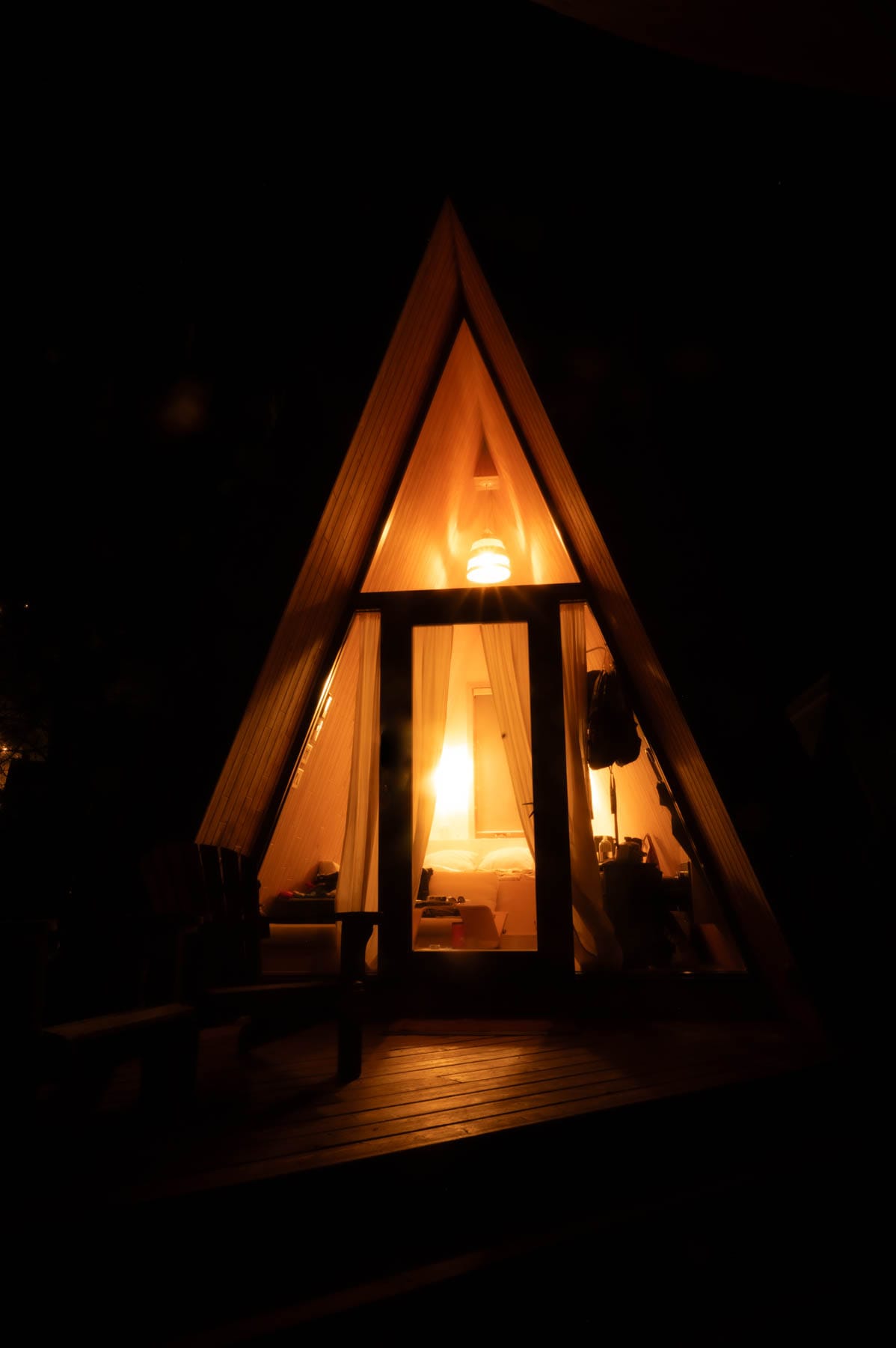 A-frame cabin at night, interior lights glowing through glass door and sheer curtains, revealing a bed and wooden deck