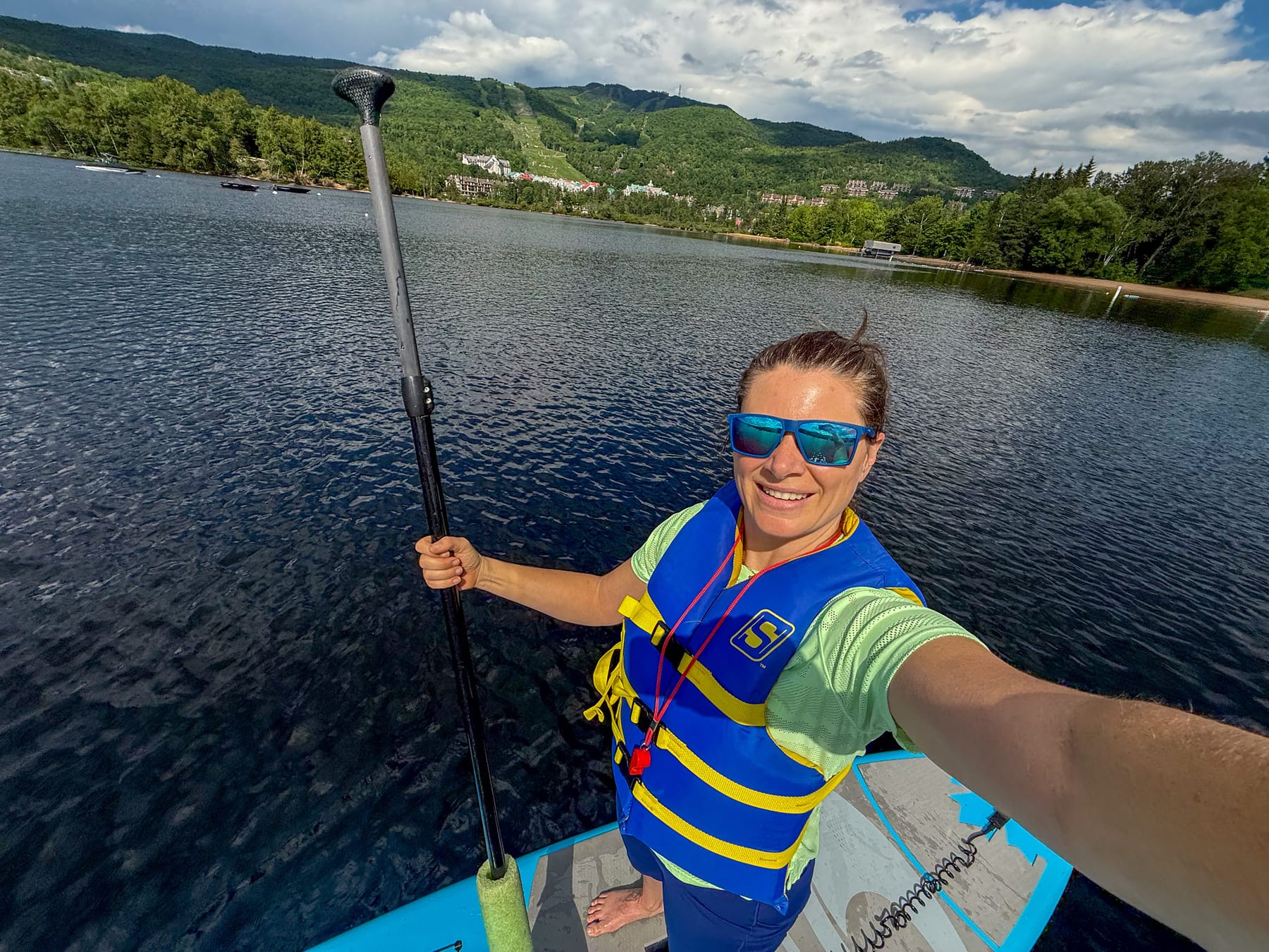 Jami Savage on paddleboard with paddle, wearing sunglasses and life vest, on lake with mountains and buildings in background
