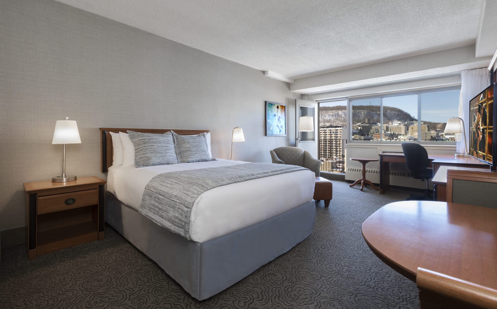 hotel room with king bed, lounge chair, desk, TV, and city view