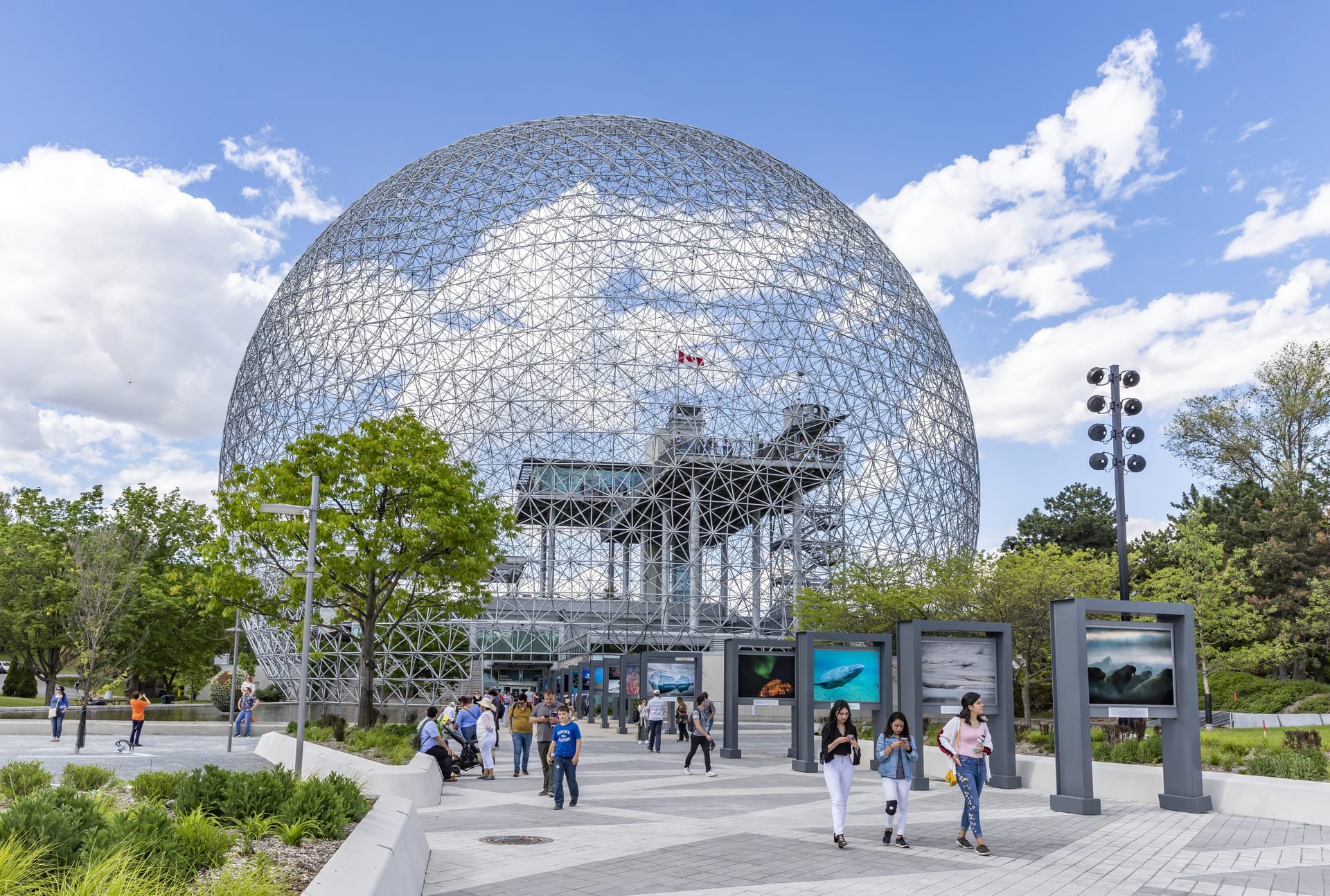The Montreal Biosphere geodesic dome with a Canadian flag inside, a paved path with people walking, trees, and outdoor exhibits