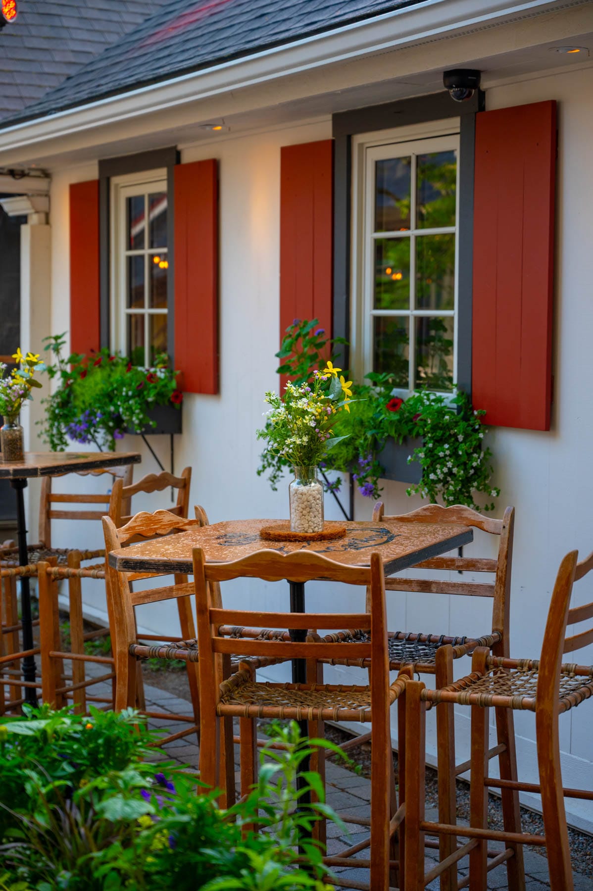 outdoor patio with wooden tables and chairs, white building, red shutters, and flower boxes with colourful blooms