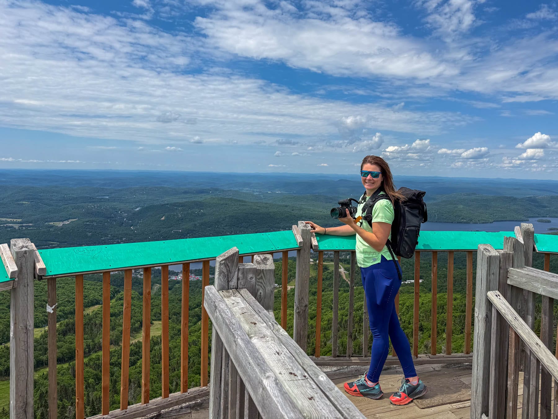 Jami Savage on observation deck holding camera, overlooking mountains, lake, and sky