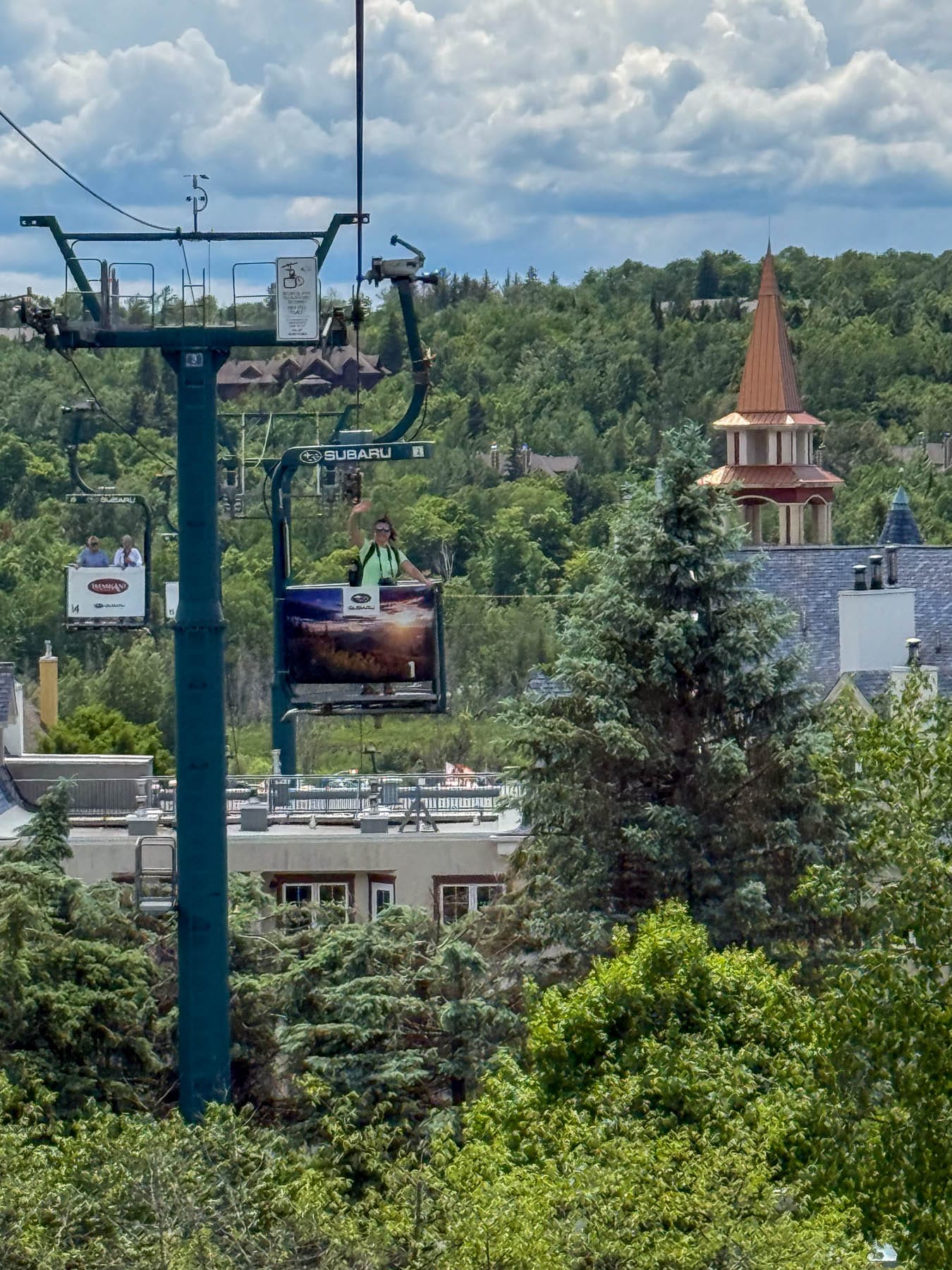 woman waving from a Cabriolet open-air lift over a treed hillside with buildings and a turret
