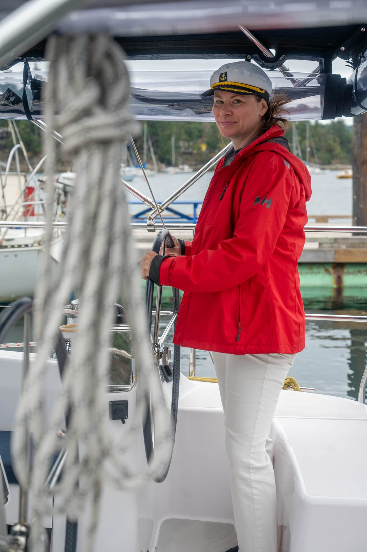 Jami Savage in red jacket and captain's hat steering a yacht in a marina, rope in foreground