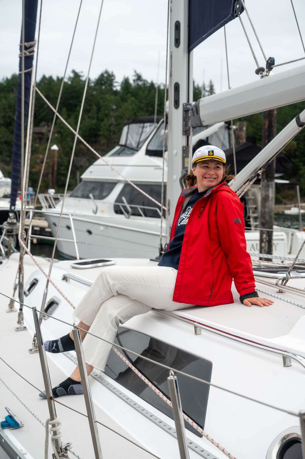 Jami Savage smiling in red jacket and captain's hat on a sailboat deck, with other boats and trees in background