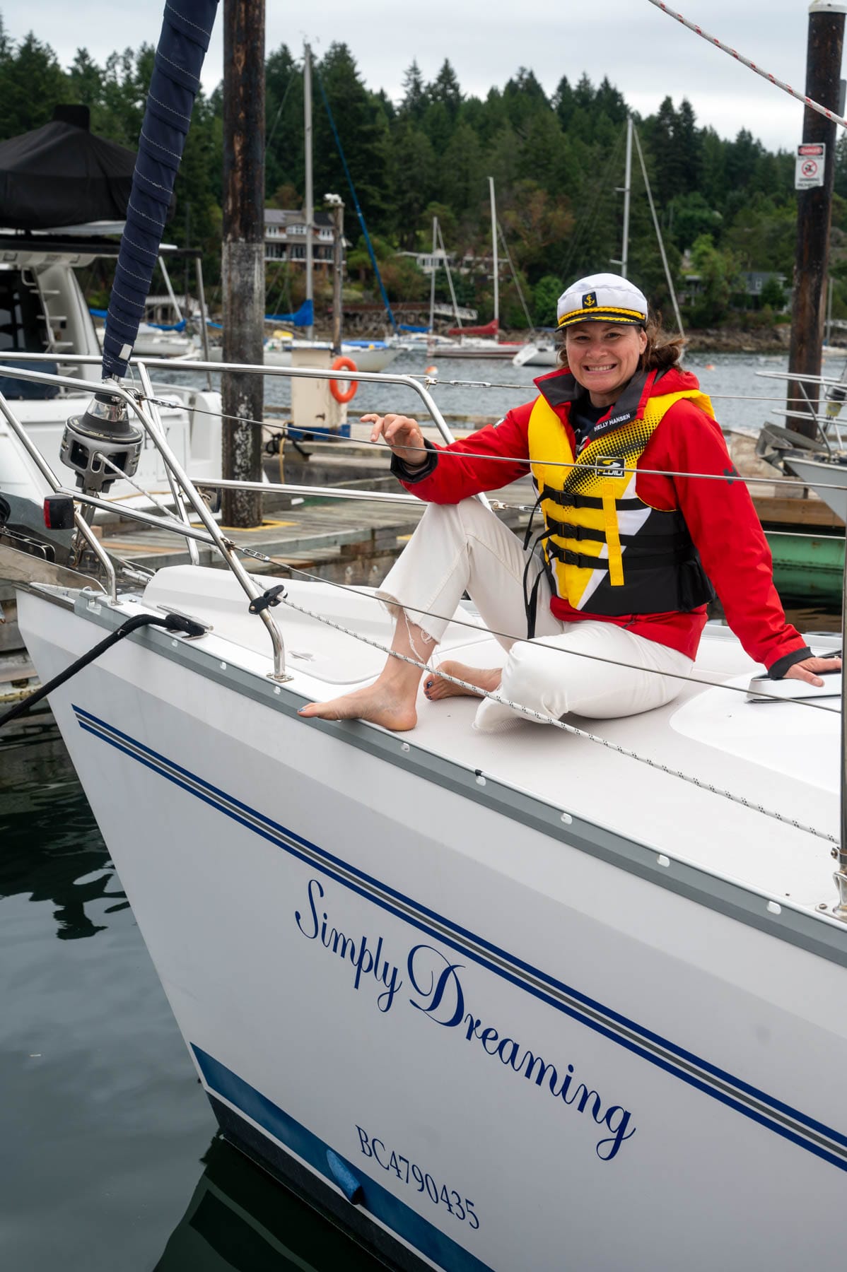 Jami Savage smiling in red jacket and life vest on a sailboat named "Simply Dreaming" at a marina