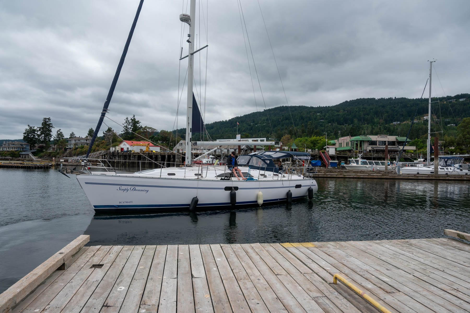 Sailboat "Simply Dreaming" approaching a wooden dock in a busy harbor with forested hills under a cloudy sky