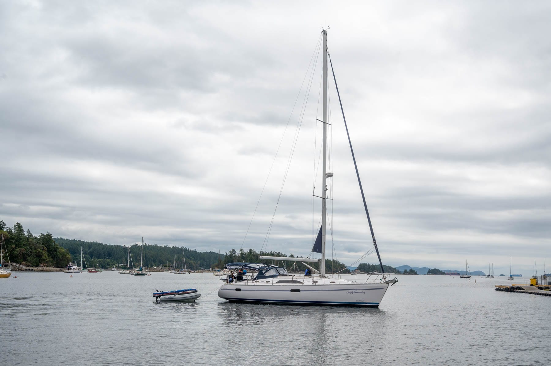 White sailboat "Simply Dreaming" with blue stripe and dinghy on grey water, cloudy sky, wooded islands and other boats in background