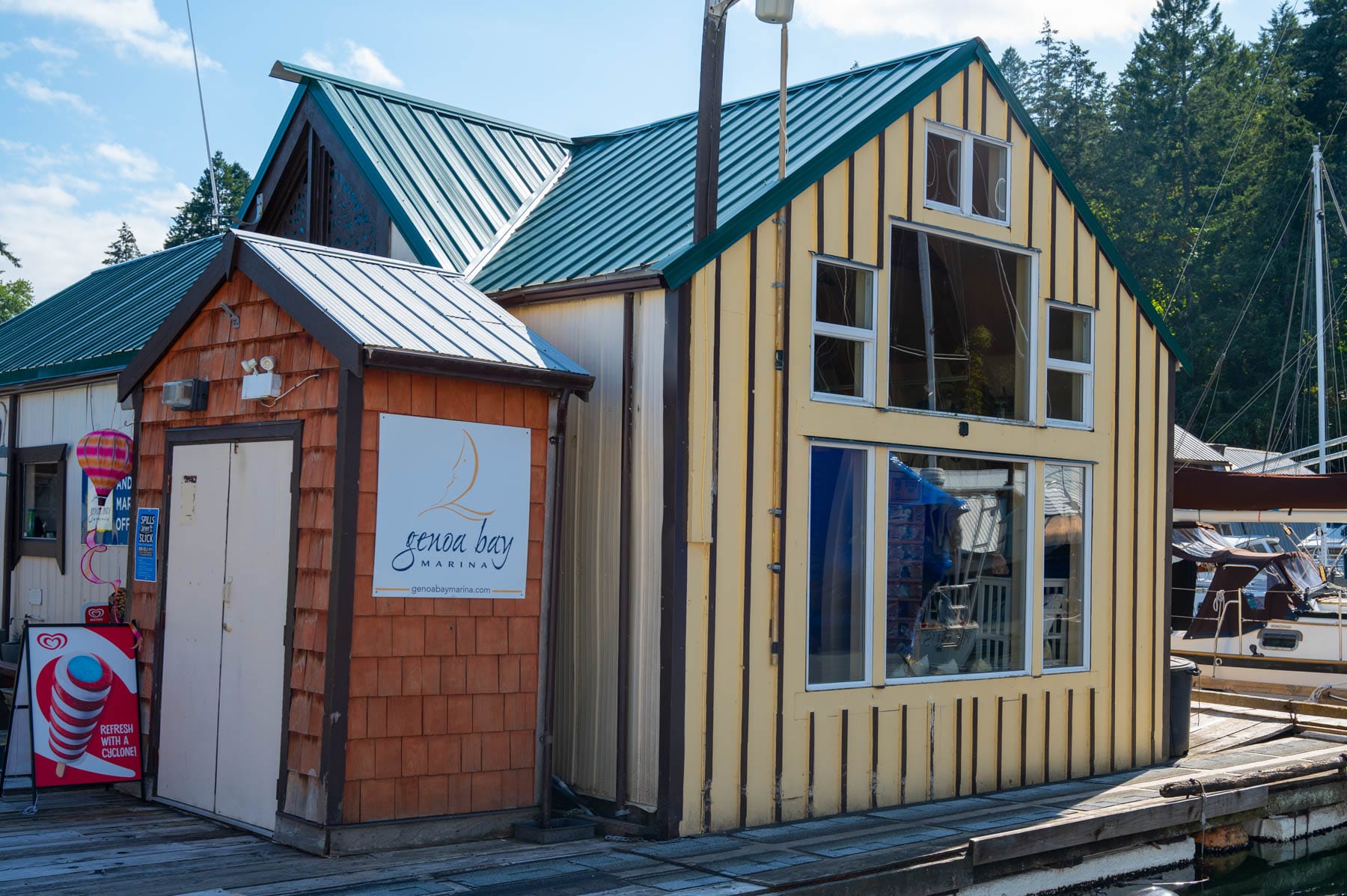 Yellow and red-shingled Genoa Bay building on a wooden dock, with a marina sign, teal roofs, boats, and trees in the background