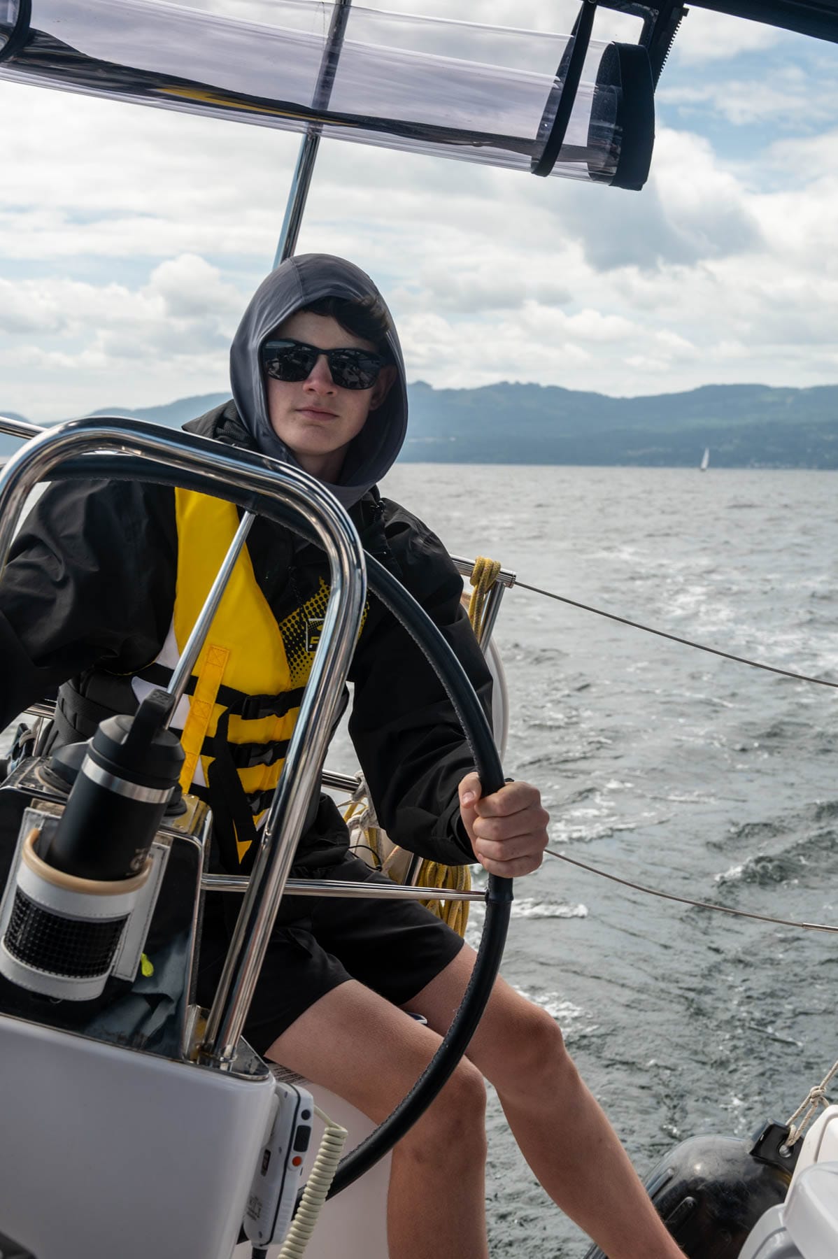 Young person in sunglasses and life vest steers a sailboat on a cloudy day with mountains in the background