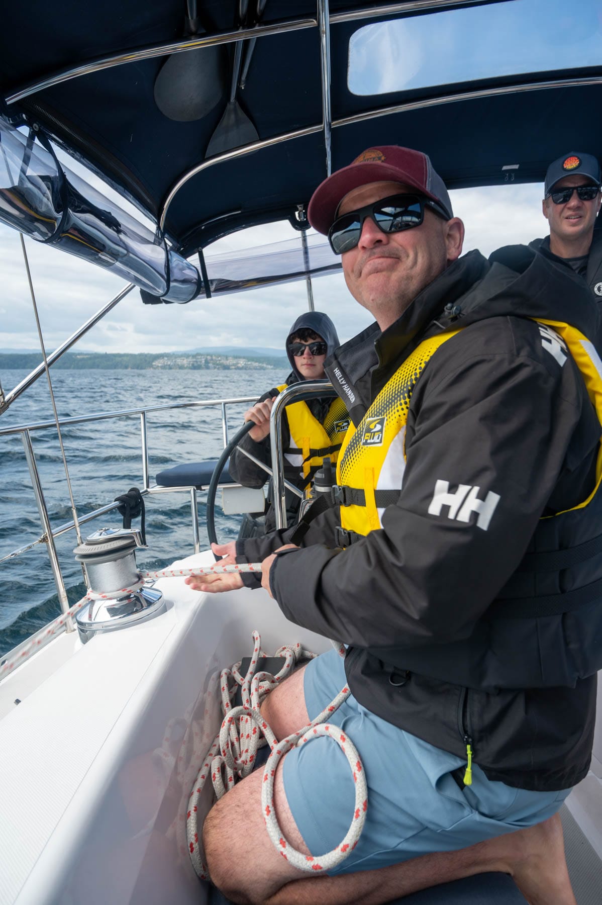 three men sailing, one working the ropes, one steering, and one in the background on a cloudy day