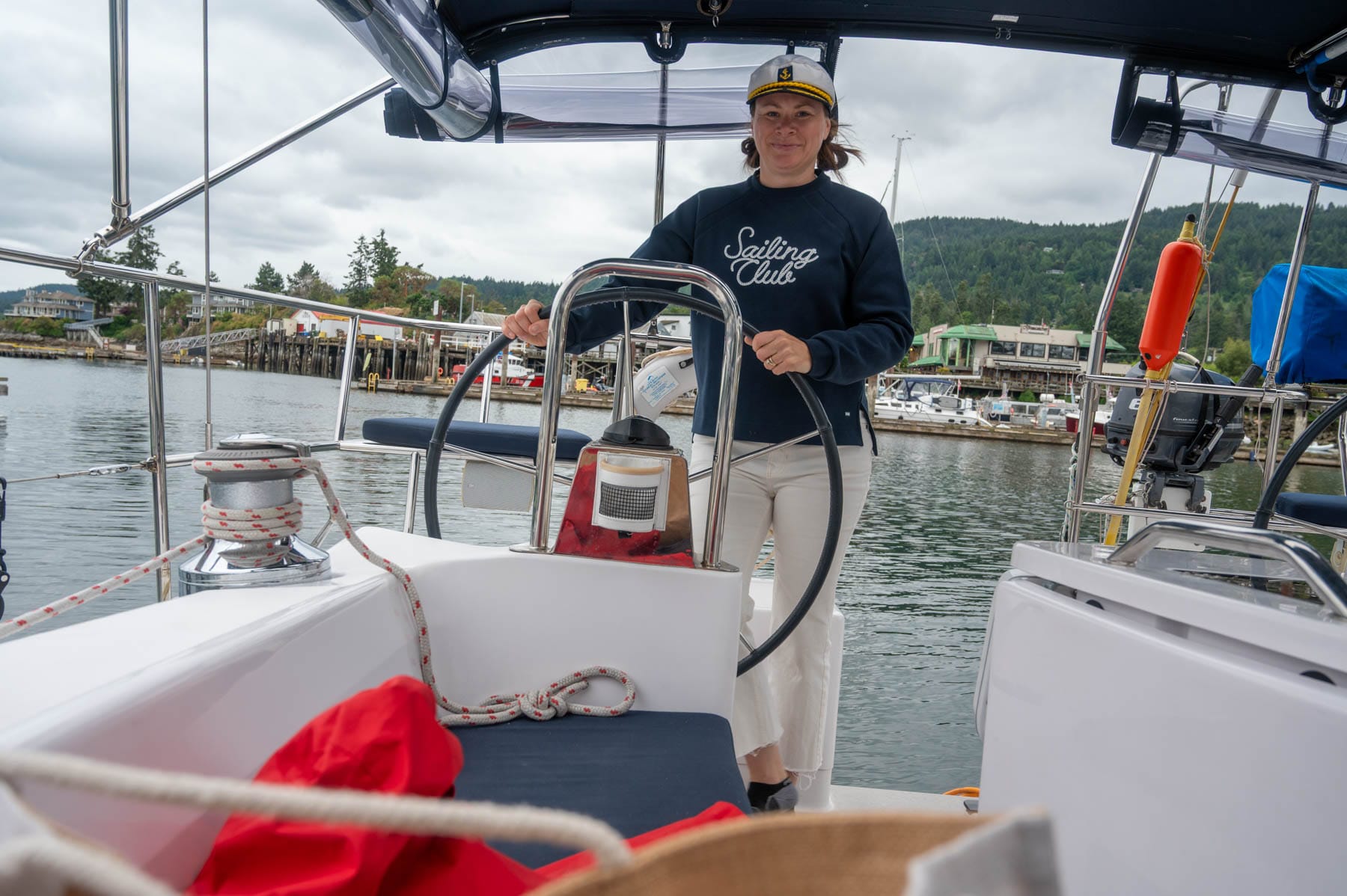 Jami Savage in captain's hat and "Sailing Club" sweatshirt steers a sailboat in a marina