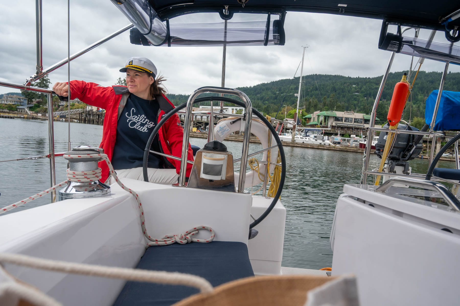 Jami Savage in red jacket and captain's hat steering a white sailboat in a harbor; other boats and town in background