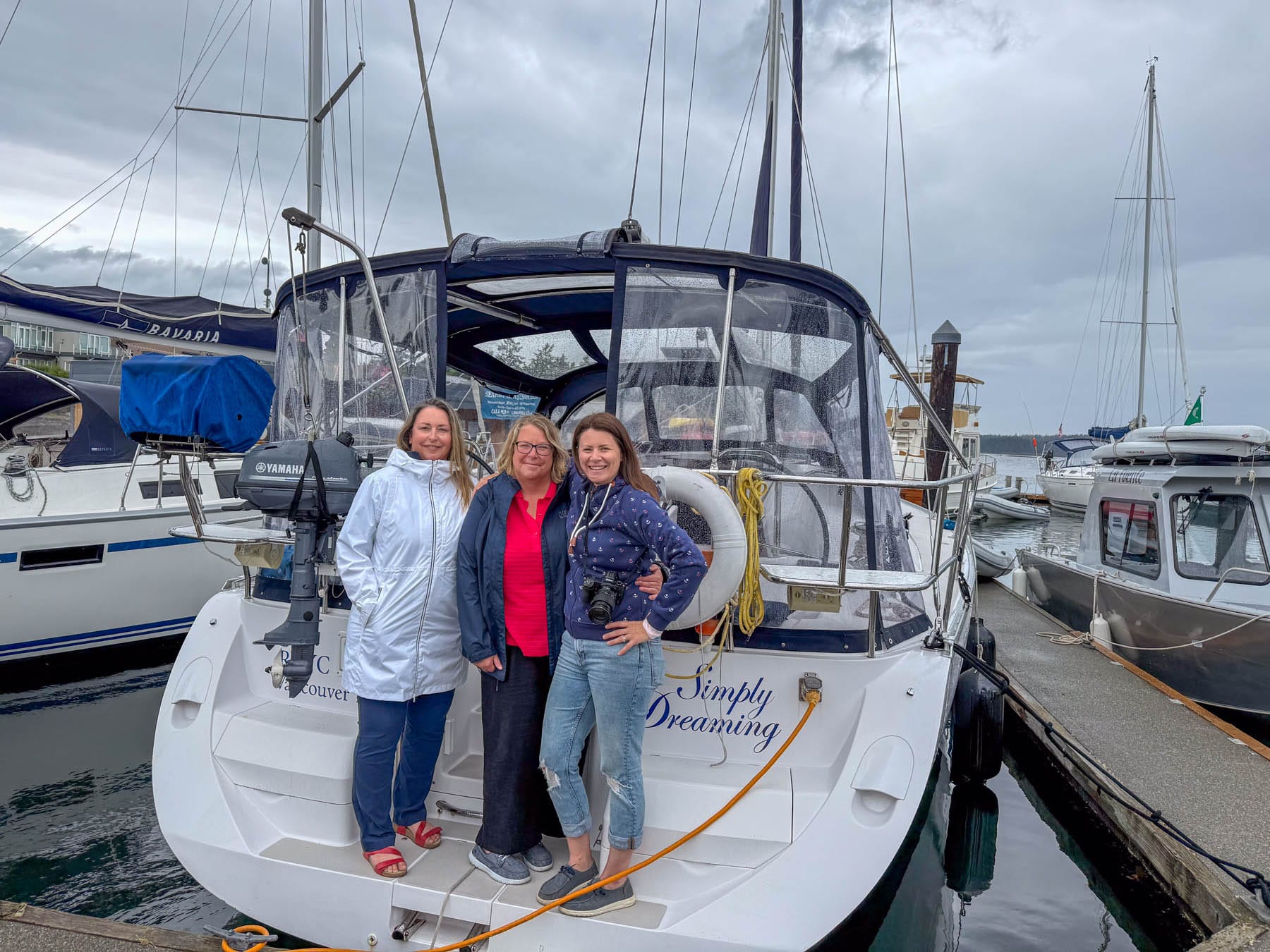 Jami Savage, Tracy Sarich, and Robin McKeown standing on sailboat "Simply Dreaming" docked in a marina under a cloudy sky, surrounded by other boats and water